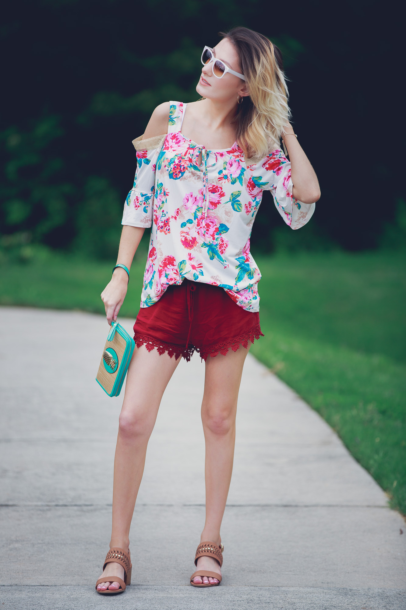 Lifestyle and fashion blogger Jessica Linn from Linn Style wearing a cold shoulder floral blouse, lace hemmed maroon shorts, low brown heels, and a teal and gold pineapple wristlet from Ross. Sunglasses from and earrings from Charming Charlie's 