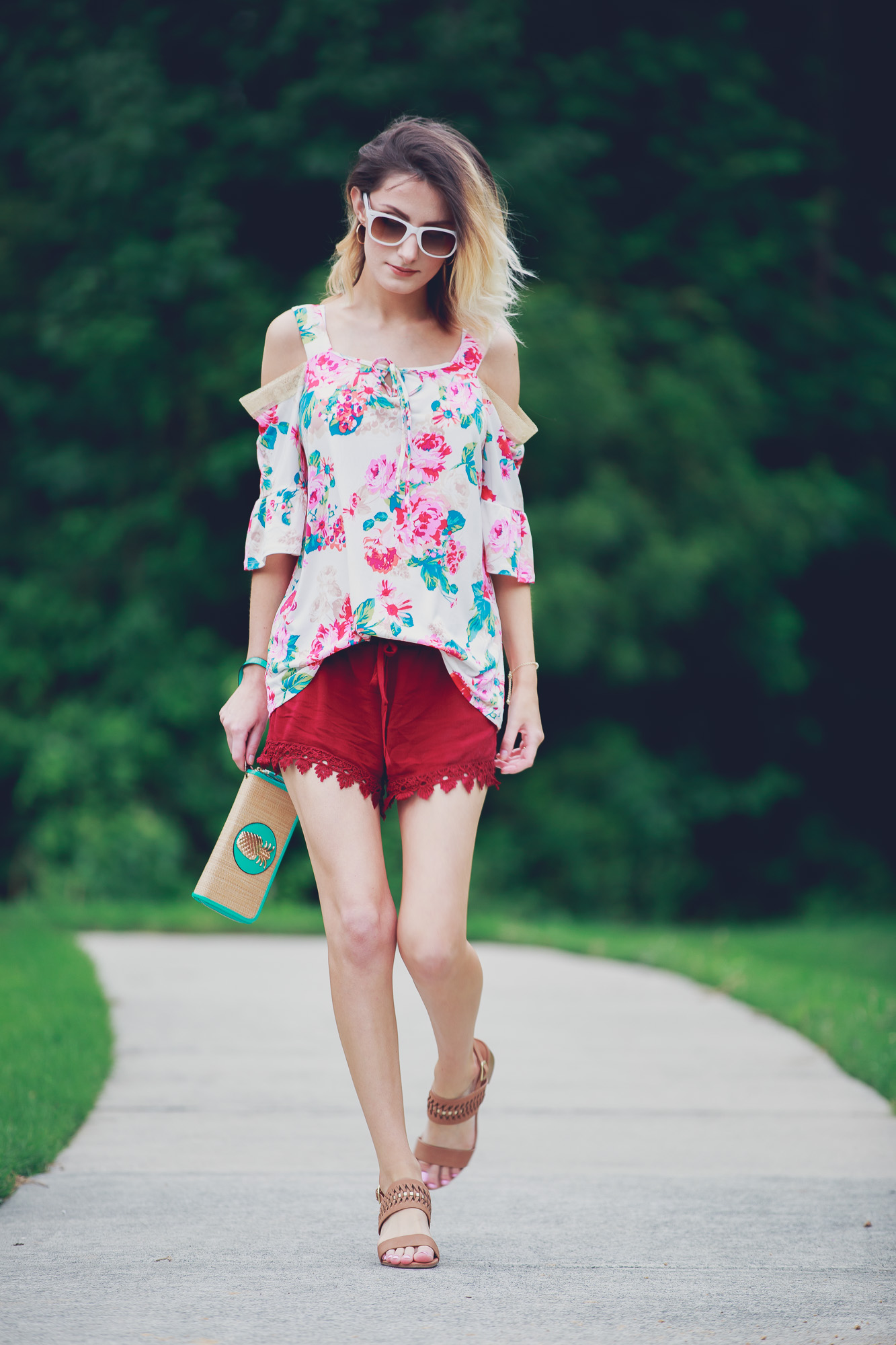 Lifestyle and fashion blogger Jessica Linn from Linn Style wearing a cold shoulder floral blouse, lace hemmed maroon shorts, low brown heels, and a teal and gold pineapple wristlet from Ross. Sunglasses from and earrings from Charming Charlie's 