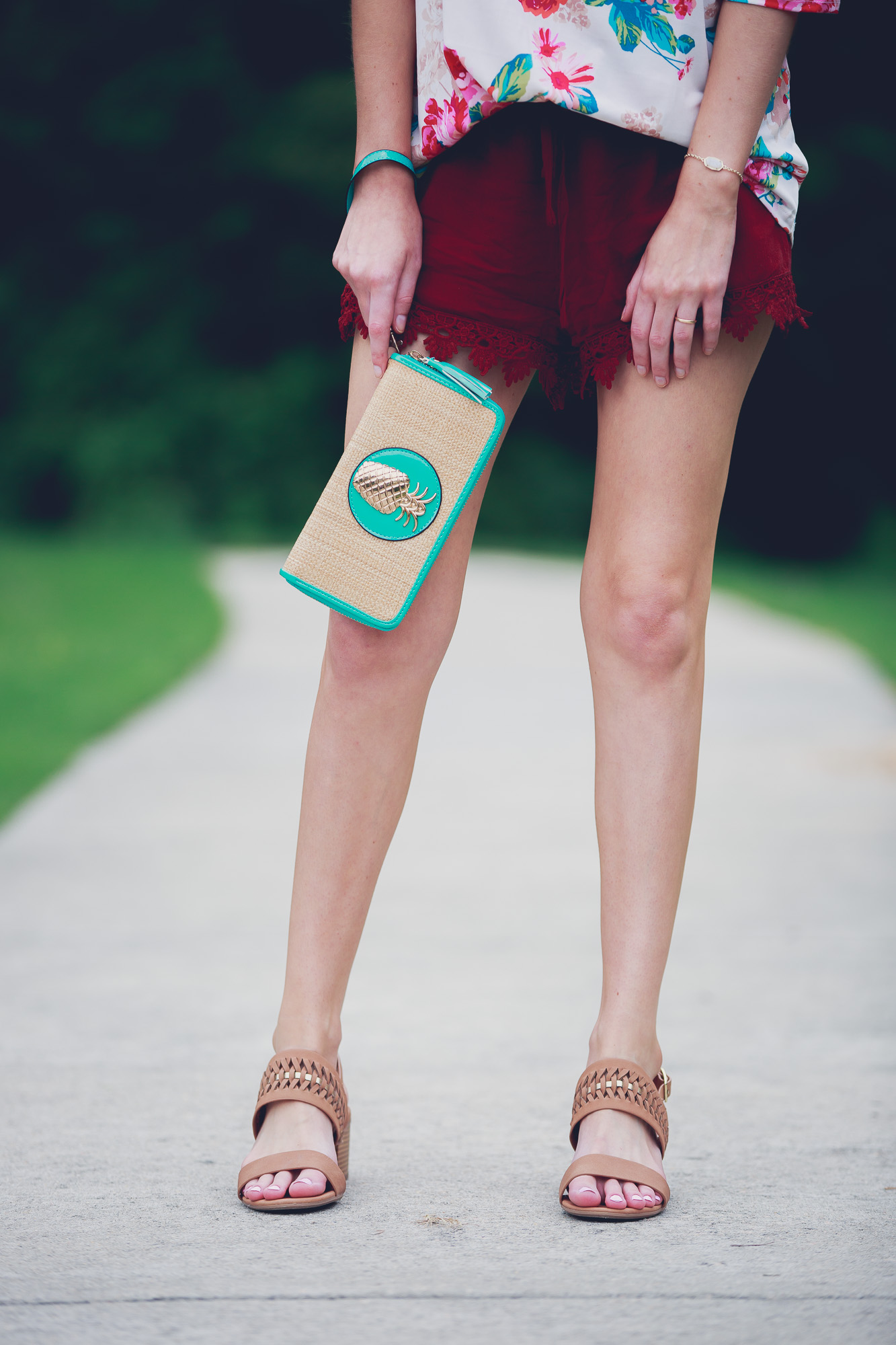 Lifestyle and fashion blogger Jessica Linn from Linn Style wearing a cold shoulder floral blouse, lace hemmed maroon shorts, low brown heels, and a teal and gold pineapple wristlet from Ross. Sunglasses from and earrings from Charming Charlie's 