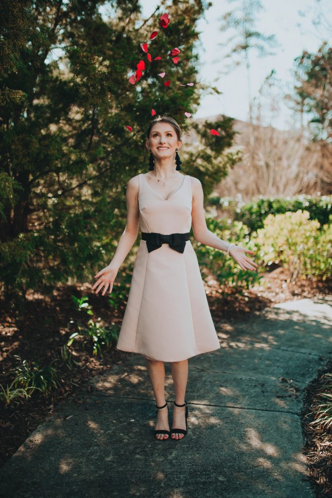 Valentines Day Outfit Inspiration | Kate Spade Fit & Flare Dress by popular North Carolina fashion and lifestyle blogger, Jessica Linn, owner and author or Linn Style and Babies, Love, & Lattes.  Jessica wearing a pale pink fit and flare dress with a black bow on the waist, black sandal heels, and throwing glitter heart confetti.