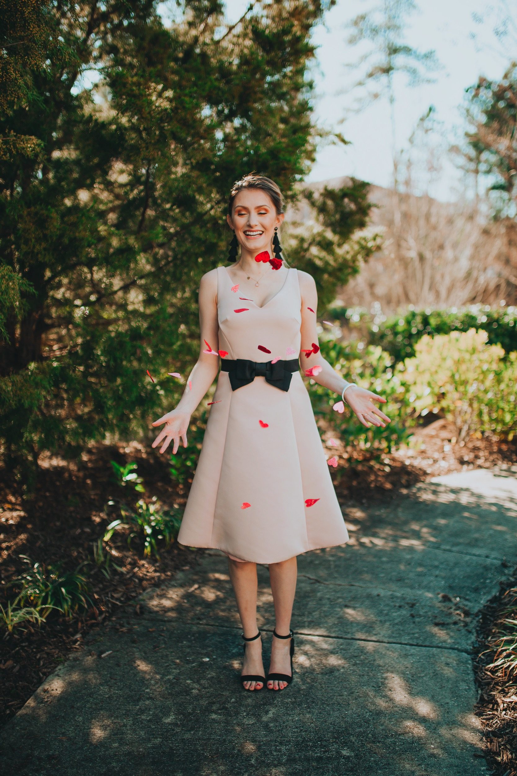 Valentines Day Outfit Inspiration | Kate Spade Fit & Flare Dress by popular North Carolina fashion and lifestyle blogger, Jessica Linn, owner and author or Linn Style and Babies, Love, & Lattes. Jessica wearing a pale pink fit and flare dress with a black bow on the waist, black sandal heels, and throwing glitter heart confetti.