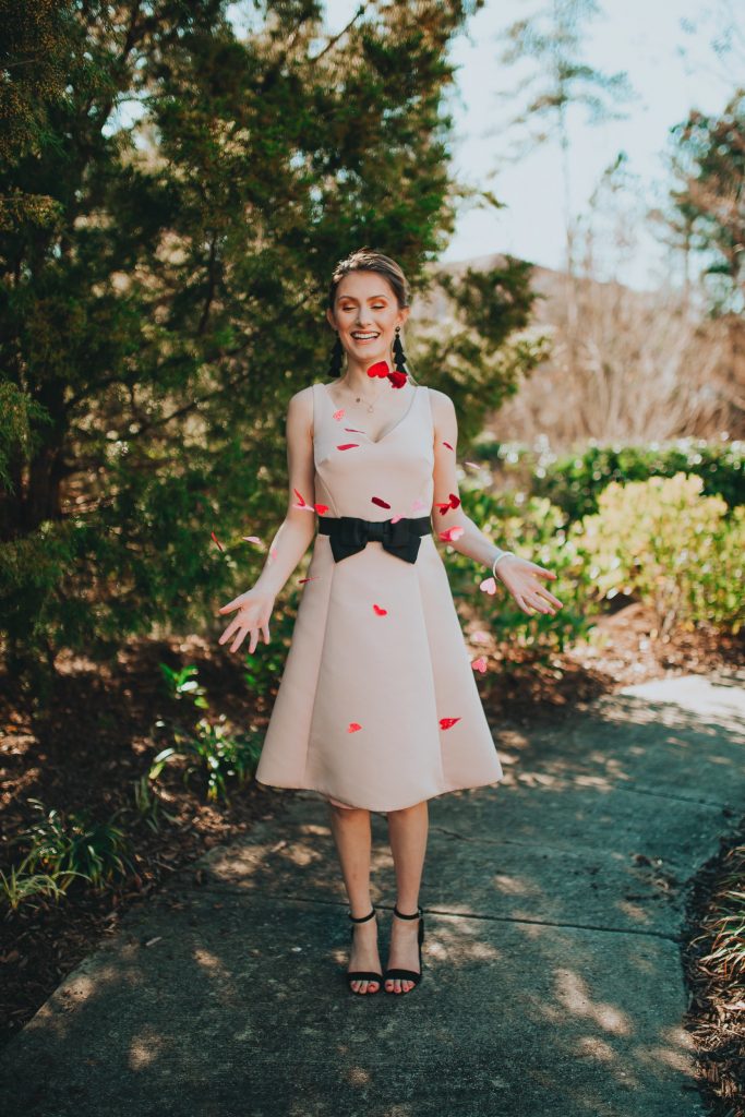 Valentines Day Outfit Inspiration | Kate Spade Fit & Flare Dress by popular North Carolina fashion and lifestyle blogger, Jessica Linn, owner and author or Linn Style and Babies, Love, & Lattes.  Jessica wearing a pale pink fit and flare dress with a black bow on the waist, black sandal heels, and throwing glitter heart confetti.